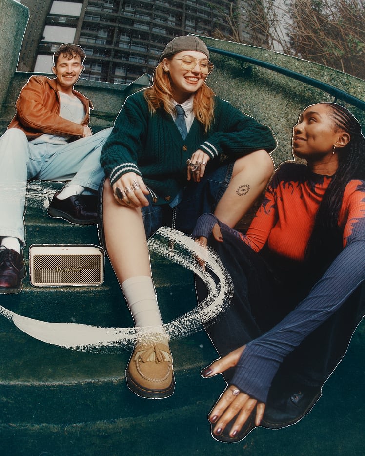 Three people sit on outdoor steps, while music plays from the Marshall Middleton II Cream portable Bluetooth speaker nearby.