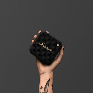 A hand with a tattoo holds a black and brass Marshall Willen II portable speaker against a plain gray background.