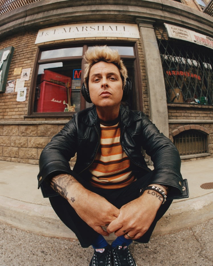 Billie Joe Armstrong con auriculares Marshall Monitor III A.N.C. Black, chaqueta de cuero y camisa de rayas sentado en la acera.