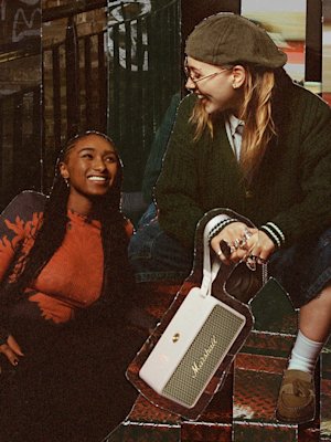 Two people sit on outdoor steps; one smiles up at the other, who wears glasses, a cap, and holds a Marshall Middleton II portable Bluetooth speaker.