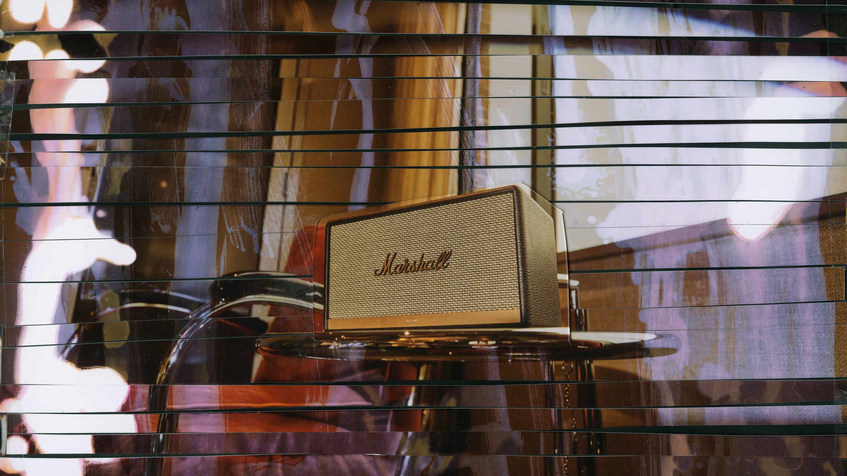 A marshall amplifier sits on a glass table with a drum set in the background, showcasing its knobs and speaker grille.
