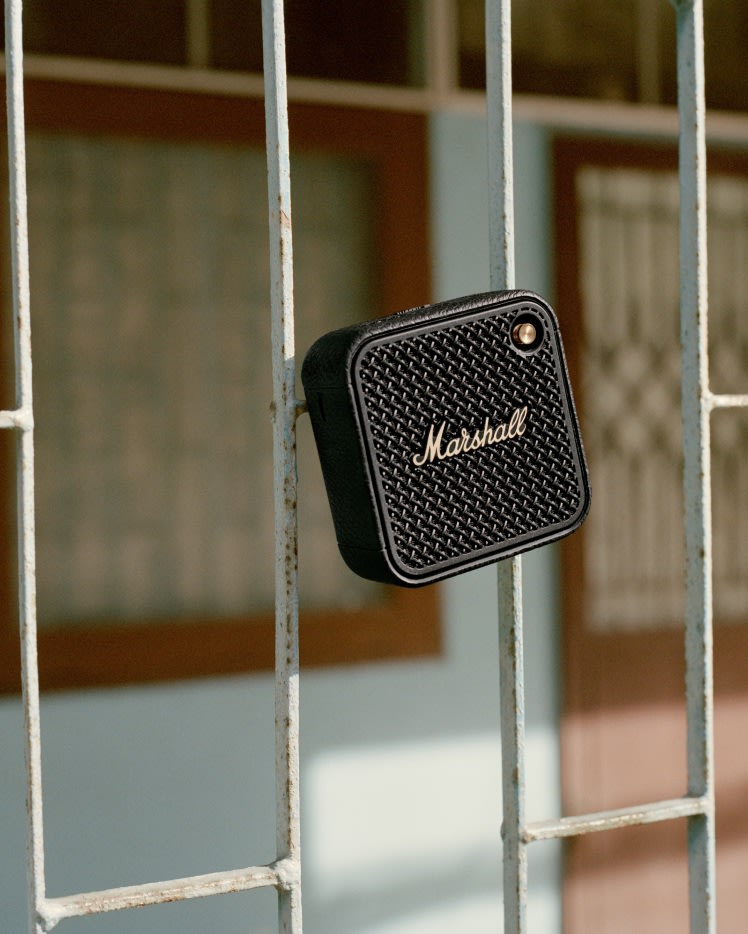 Marshall Willen II Black and Brass portable speaker with the iconic logo is hanging on a metal fence.