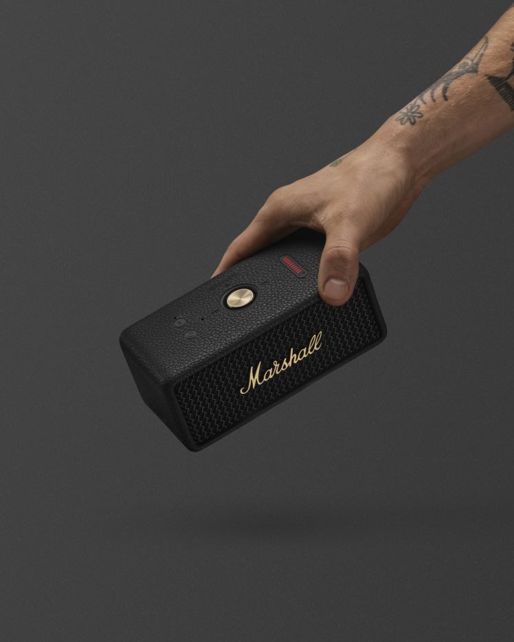 A hand holding Marshall Emberton III Black and Brass bluetooth speaker against a dark gray background.