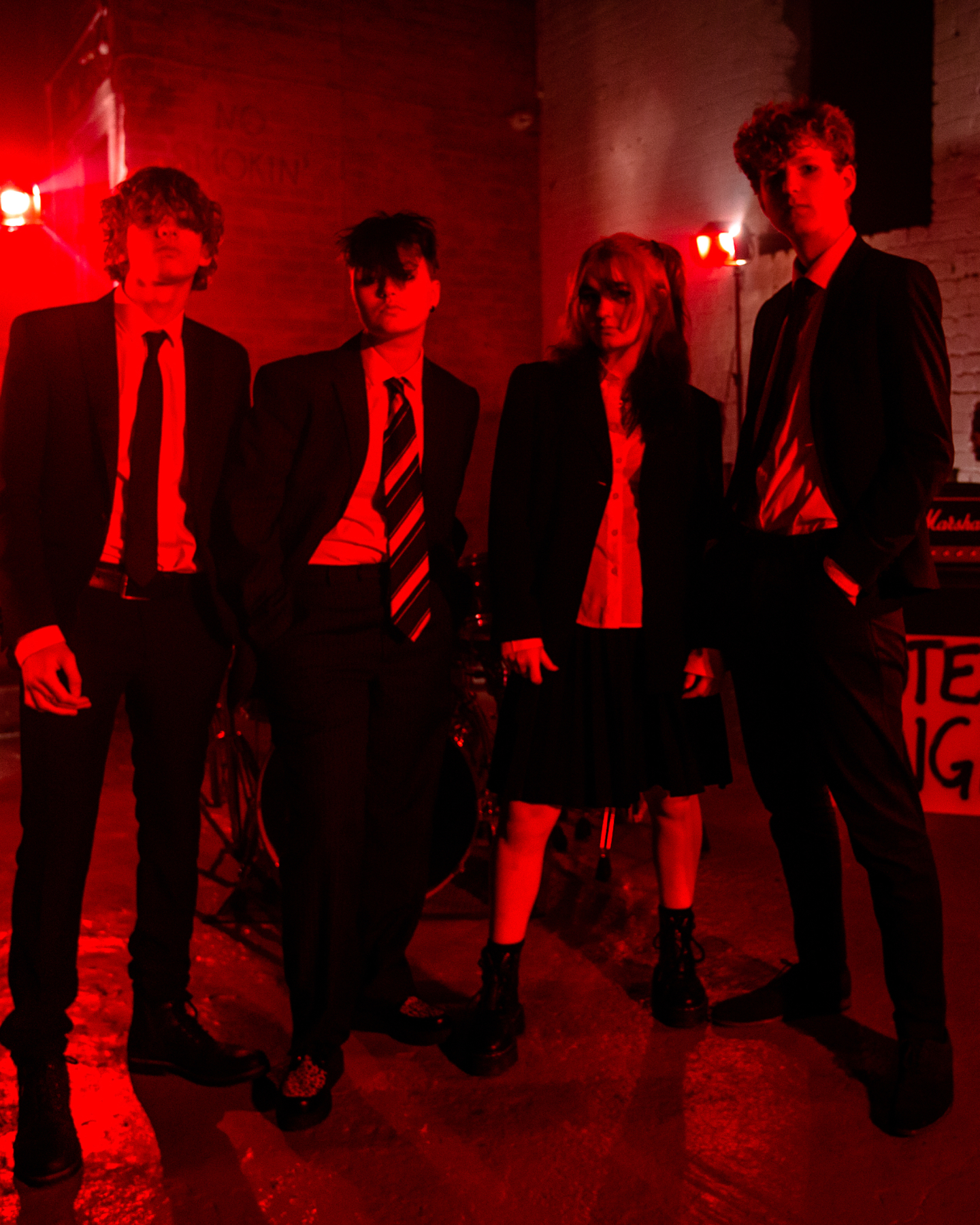 Band Noah and the loners stand in a dimly lit red room, dressed in formal attire. There is a drum set in the background.