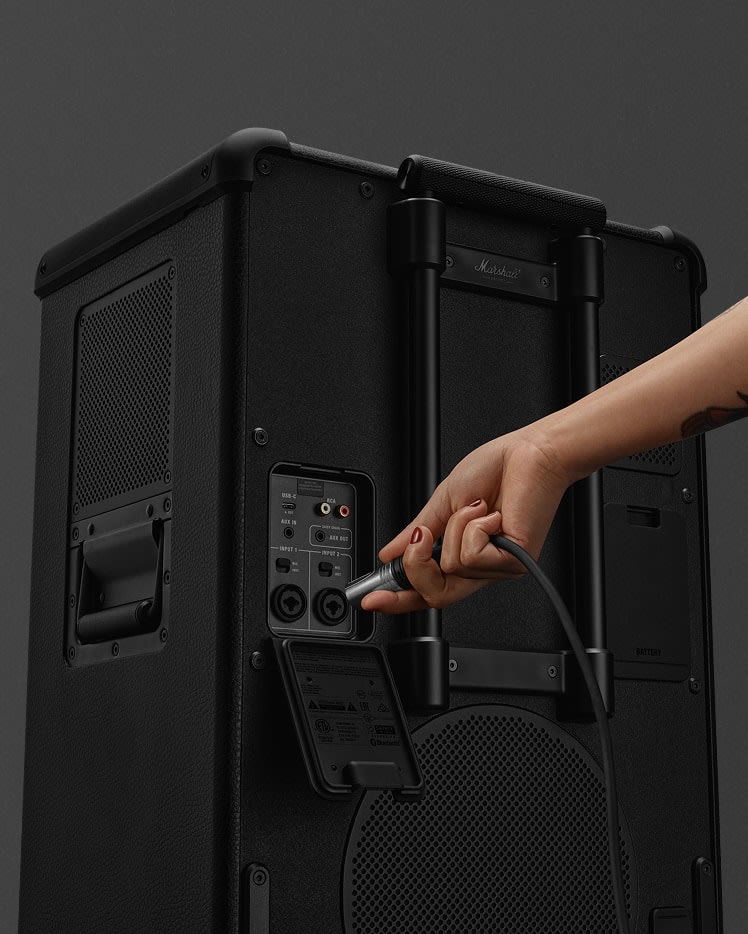 A hand plugs a mic cable into the input panel of a black Marshall Bromley 750 portable party speaker.