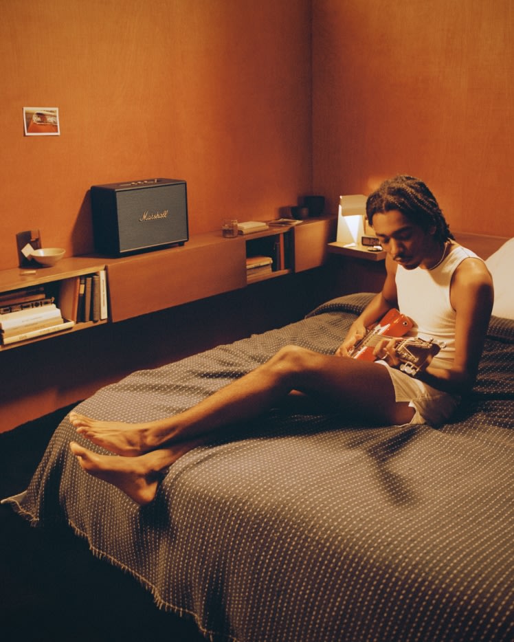 Ein Mann sitzt auf einem Bett mit einer Marshall WOBURN III BLACK Gitarre.