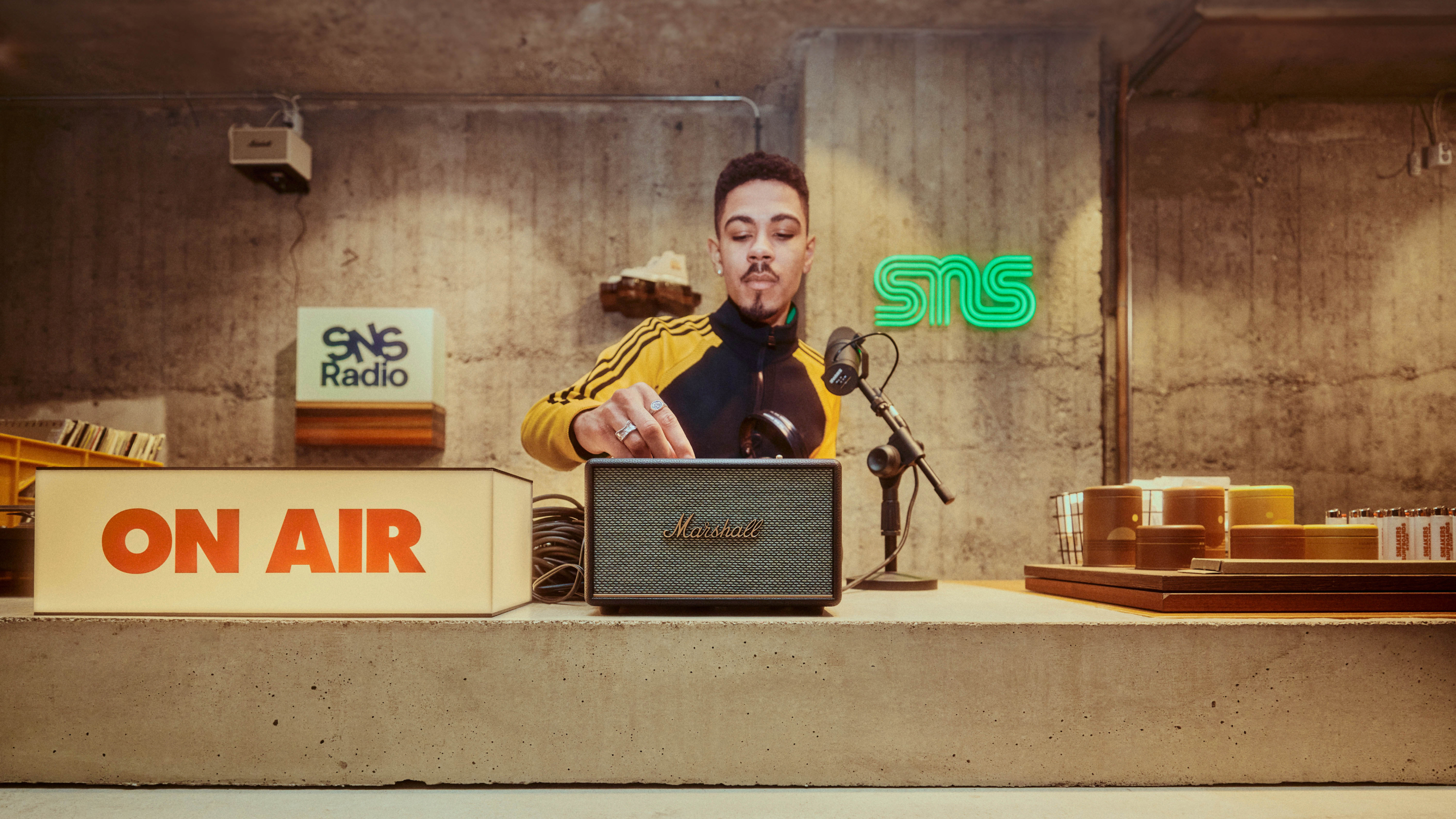 Ein junger Mann stellt die Regler eines Marshall-Lautsprechers in einem Radiosender mit Neonschildern und Vintage-Dekor ein.