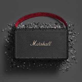 Marshall Kilburn III Black and Brass portable speaker, featuring a red handle, sits on a dark surface with scattered black stones, emphasizing its sleek and stylish design.