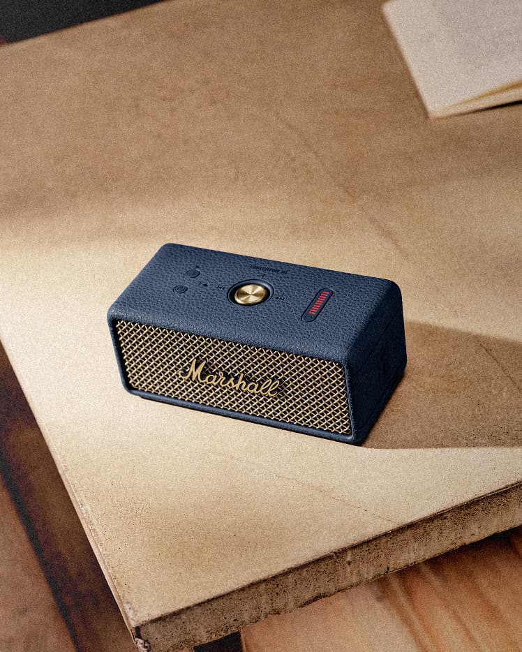 Marshall Emberton III Midnight Blue portable speaker sits on a wooden surface near a stack of papers, delivering powerful Bluetooth sound.