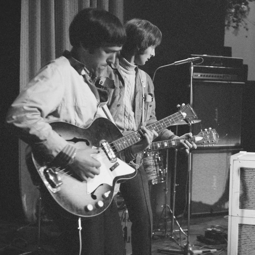 Fotografía en blanco y negro de un grupo de personas tocando la guitarra con amplificadores Marshall de fondo.