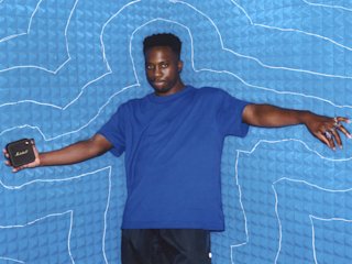Kelvin Krash in blue shirt holding a Marshall Willen portable bluetooth speaker against a blue patterned background.