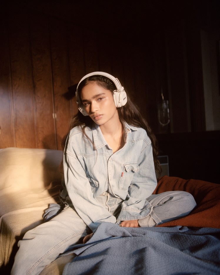 Eine Frau, die Jeanskleidung und Marshall Major V Cream On-Ear-Kopfhörer trägt, sitzt auf einem Sofa.