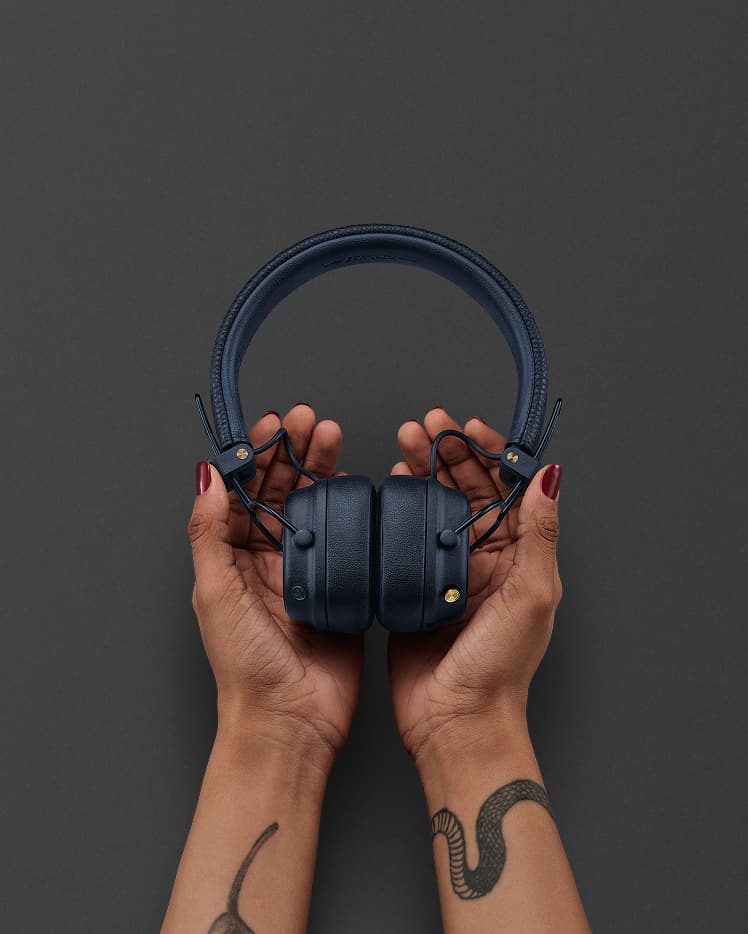 Tätowierte Hände halten ein Paar Marshall Major V Midnight Blue Kopfhörer vor einem schlichten dunklen Hintergrund.