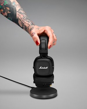 A tattooed hand placing a pair of Major IV headphones with the right ear cup down on a wireless charging pad.