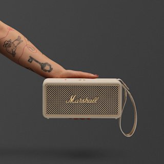 A hand with tattoos holds a Marshall Middleton II portable Bluetooth speaker in Cream with a wrist strap against a plain gray background.