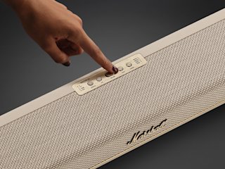 A hand presses a button on the control panel of the Marshall Heston 60 Cream soundbar speaker against a dark background.