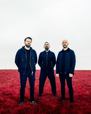Three men stand on a field of red grass under a cloudy sky, all wearing dark clothing.