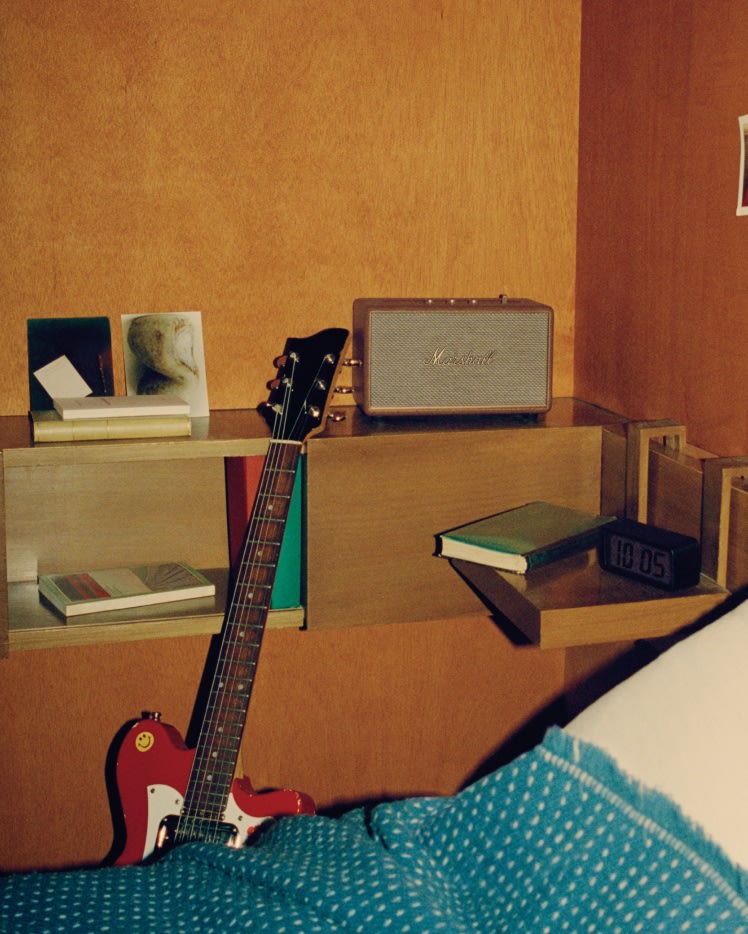 A brown speaker, known as the Marshall Acton III Brown, sits on a bed alongside a guitar.