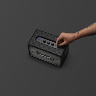 A hand adjusts a control knob on a black checkered Marshall Acton III Awake NY speaker placed on a dark surface.