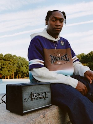 A person sits outdoors next to a Marshall Acton III Awake NY Edition speaker, with blue sky in the background.