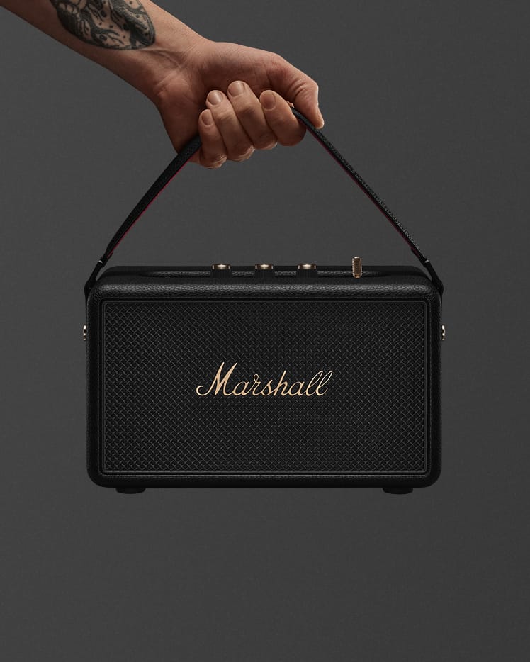 A tattooed hand holds the Marshall Kilburn III Black and Brass portable speaker by its handle against a plain background.