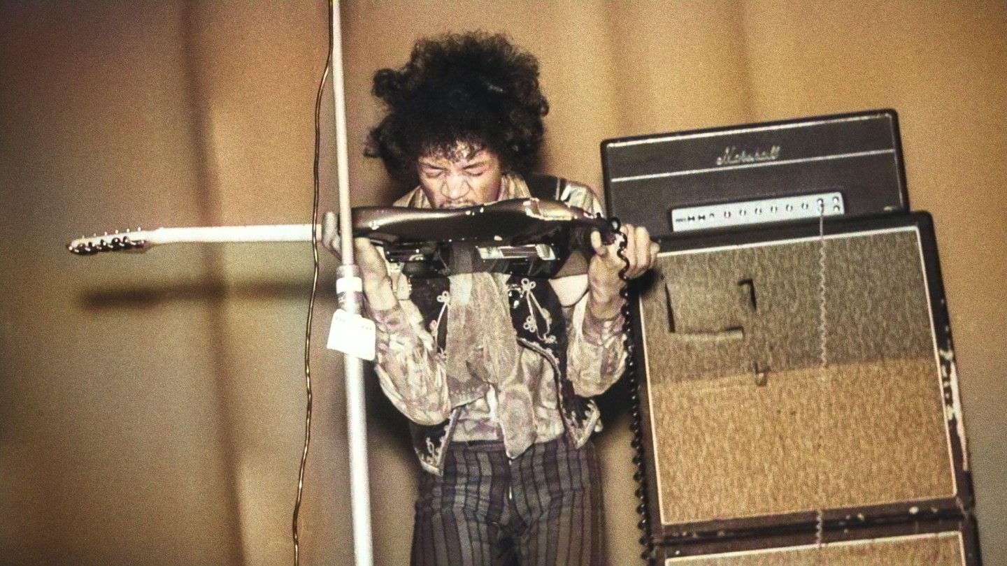 Jimi Hendrix spielt auf der Bühne eine Gitarre mit den Zähnen, trägt ein geblümtes Hemd und eine gestreifte Hose und steht neben einem Verstärker.