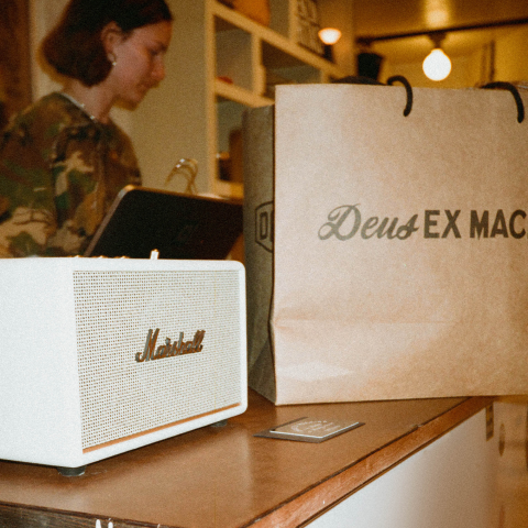 A woman works on a laptop behind a counter near a marshall speaker and a deus ex machina branded shopping bag.