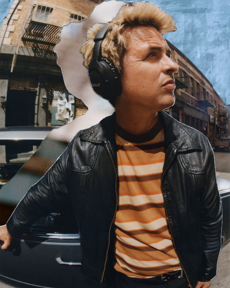 Billie Joe Armstrong waering Marshall Monitor III A.N.C. Black over-ear headphones, a striped shirt, and a leather jacket stands next to a car on a street, looking upwards.