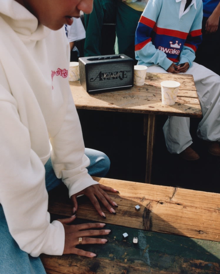 A person in a white hoodie sits on a wooden bench with Acton III Awake NY Edition speaker by Marshall and paper cups in the background.