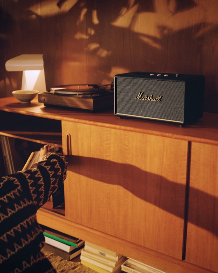 A man is sitting in front of a bookcase with a Marshall Stanmore III Black speaker.