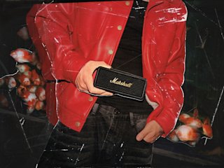 A man in a red jacket holding a Marshall Emberton III Black and Brass portable bluetooth speaker.