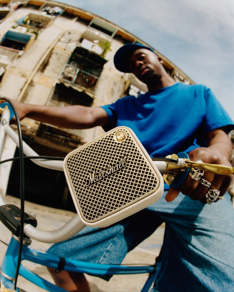 A man in a blue shirt and cap rides a bicycle with a portable Marshall Willen II bluetooth speaker.