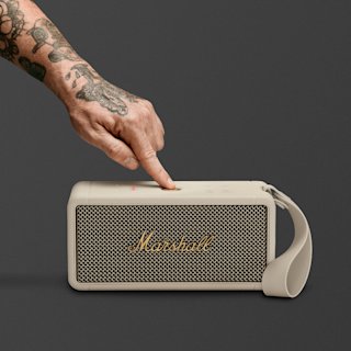 A tattooed hand presses the control button on the Cream Marshall Middleton portable speaker with a carrying strap, against a plain dark background.