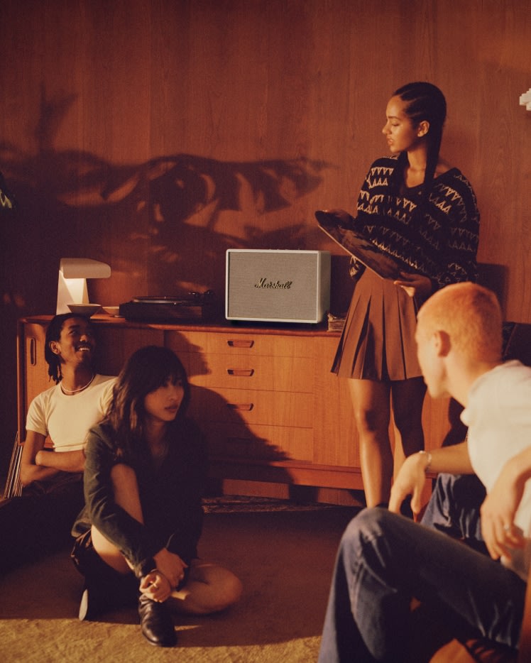 A group of people sitting on the floor in front of a white Marshall WOBURN III CREAM speaker, watching television.