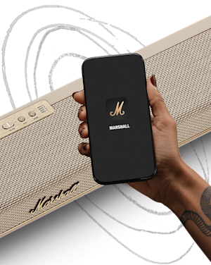 A hand holds a smartphone displaying the Marshall app in front of a Marshall Heston 60 Cream soundbar speaker with visible control buttons and logo.