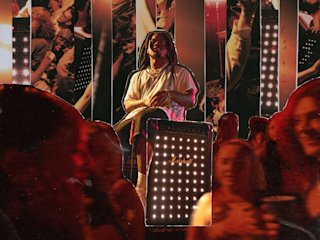 A man sits on a stool behind a Marshall Bromley 750 party speaker, with a crowd and vibrant concert lighting in the background.