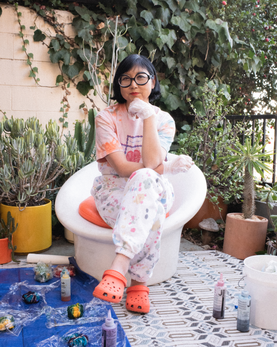 Woman with glasses and colorful attire sitting in a white chair surrounded by paint supplies and plants in a courtyard.