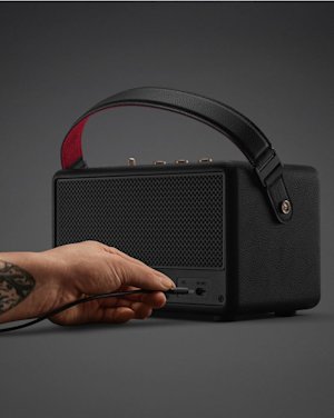 A hand plugs a cable into the input port of the Marshall Kilburn III Black and Brass portable speaker, featuring a textured finish and a leather handle.