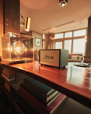 Ein Marshall-Lautsprecher auf einem Holztisch in einem gemütlichen, warm beleuchteten Wohnzimmer mit moderner Einrichtung und großen Fenstern.