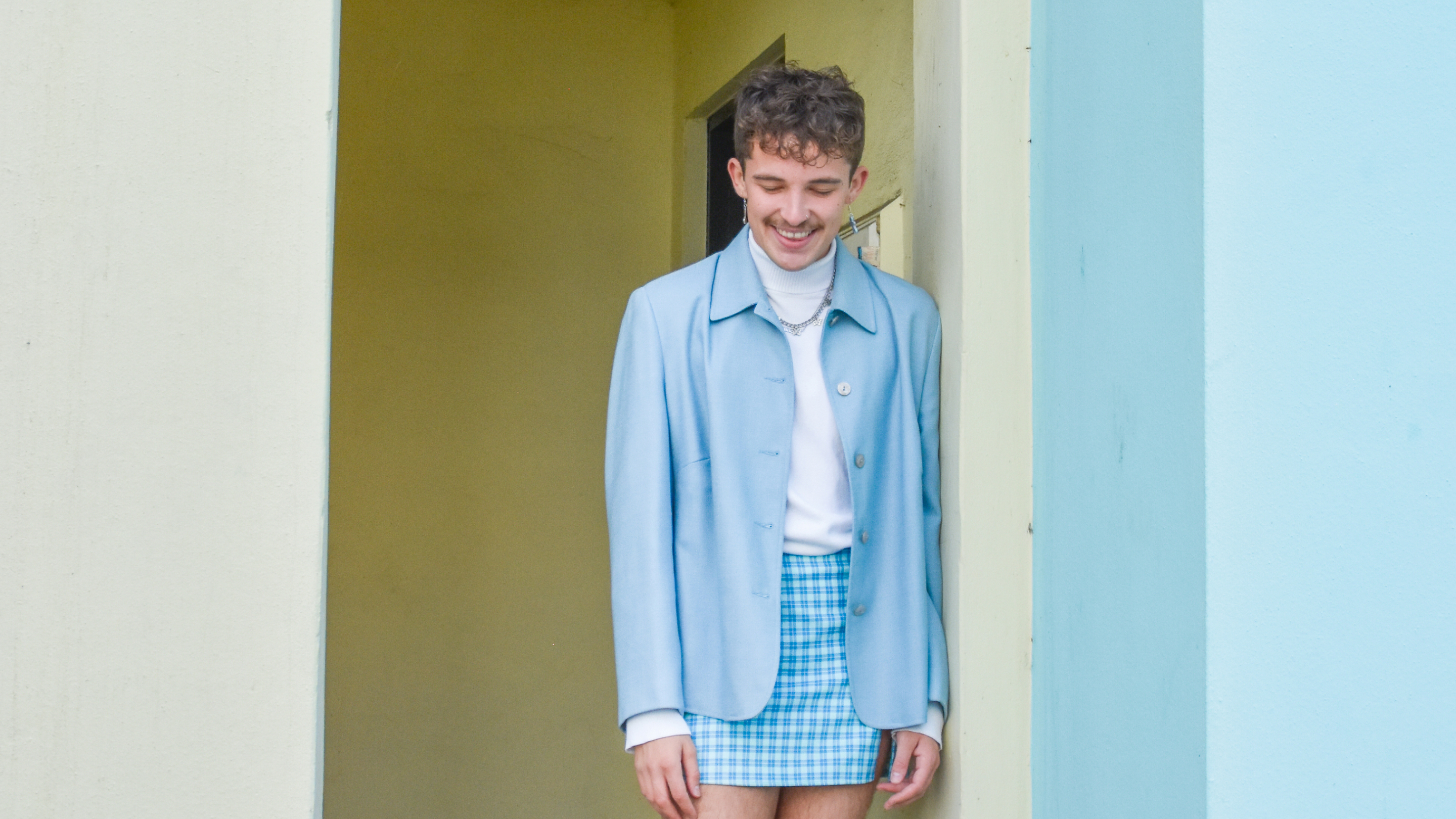 Freddie Lewis in a blue set of Jacket and skirt standing against a yellow wall. 