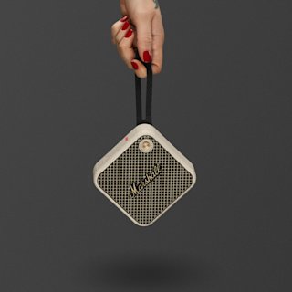 A hand with red nails holds a Marshall Willen II portable speaker in cream by its strap against a plain dark background.