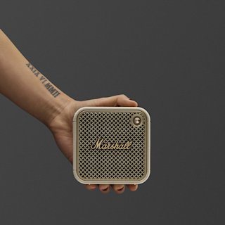 A hand with red nails holds a Marshall Willen II portable speaker in cream by its strap against a plain dark background.