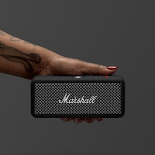 A hand with red nail polish holds a Marshall Emberton II portable speaker in Black and Steel against a plain dark background.