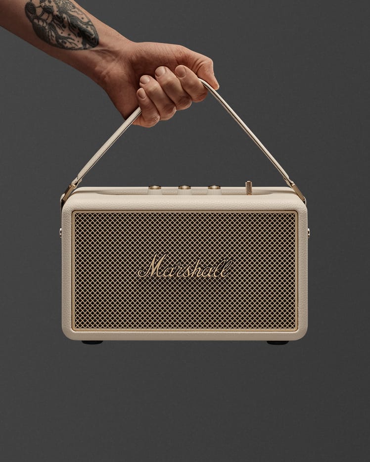 A hand holds a Marshall Kilburn III Cream portable speaker by its strap against a plain green background.