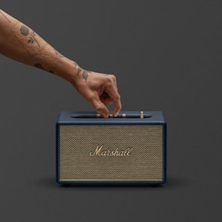 A tattooed hand adjusts a knob on a Midnight blue Marshall Acton III speaker, set against a plain dark background.