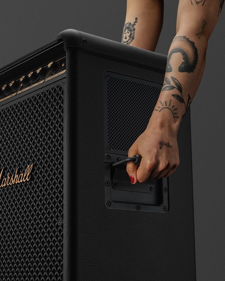 A tattooed arm grips the handle of a large black Marshall Bromley 750 speaker amplifier.