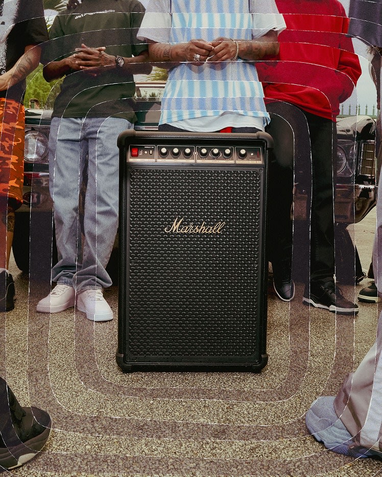 Five people stand behind a large Marshall Bromley 750 party speaker on the pavement, with part of a car visible in the background.
