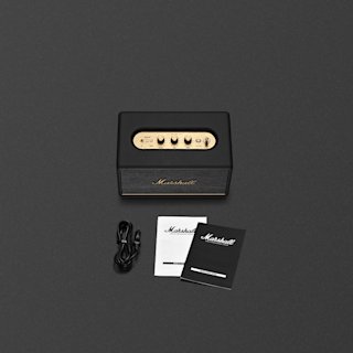 Marshall Acton III home speaker with brass control panel, power cord, user manual, and warranty booklet displayed on a dark surface.