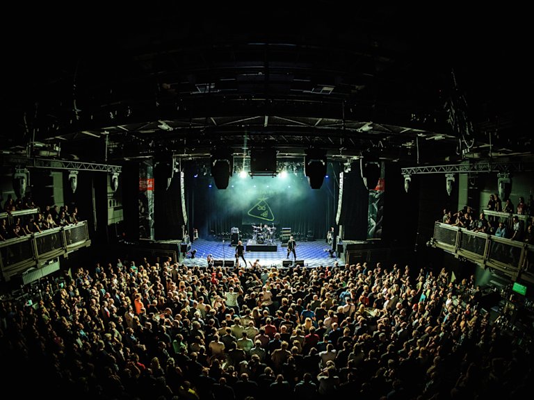 A large crowd watches a band perform on stage in an indoor concert venue with balcony seating.