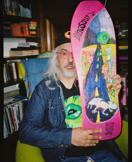 J Mascis sitting in a chair and holding a skateboard.